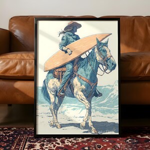 May include: Framed art print depicting a person riding a horse on a beach, carrying a surfboard. The artwork features a color palette of blue, tan, and white. The horse is blue and white, and the person is wearing a hat and denim clothing. The background shows ocean waves.