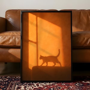May include: A rectangular wall art print in a thin black frame featuring a warm orange-toned image of a cat silhouette walking across a surface, with a vertical window shadow cast across the background, creating a high-contrast visual effect.