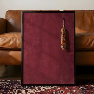 May include: A rectangular wall art piece featuring a deep burgundy velvet-textured background enclosed in a thin black frame. A small, detailed figurine of a tiger is attached to the upper right side, hanging vertically with its head facing downward.