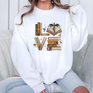 May include: White sweatshirt with a colorful graphic design. The design spells out "LOVE" using stacks of books and an open book with a dragon. Small dragons and stars are scattered around the letters.