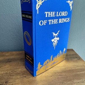 Puede incluir: Un libro de tapa dura azul titulado "THE LORD OF THE RINGS" con letras doradas y detalles decorativos. El lomo dice "ONE VOLUME" y "LORD OF THE RINGS". El libro está sobre una superficie de madera.