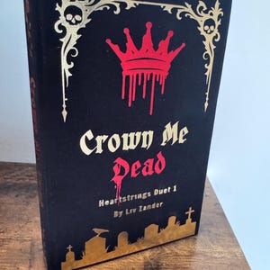May include: A black hardcover book titled Crown Me Dead by Liv Zander, Heartstrings Duet 1. The cover features a red dripping crown, gold filigree corners with skulls, and a gold silhouette of a graveyard with tombstones and a bird at the base.