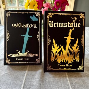 Op de afbeelding: Twee hardcover boeken met zwarte covers en gouden letters. Het boek links heeft de titel "Quicksilver" met een blauwe zwaardillustratie. Het boek rechts heeft de titel "Brimstone" met een gouden vlamillustratie. Beide boeken zijn van Callie Hart.