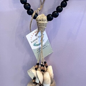 May include: A decorative wall hanging featuring a strand of black beads, a woven jute element, and a cascade of seashells. The hanging includes a tag with the text "Lunar Tides". The seashells are white and beige.