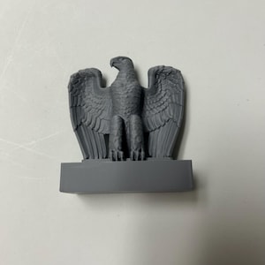 May include: A matte gray eagle figurine standing on a rectangular base. The eagle is depicted with its wings spread wide, showing detailed feather textures on the wings and body. The bird faces toward the left with a sharp, hooked beak.