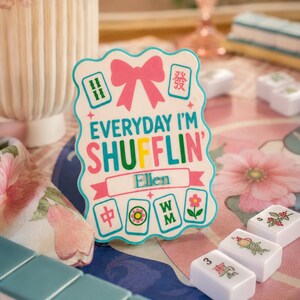 Może przedstawiać: Biały znak z napisem "EVERYDAY I'M SHUFFLIN' Ellen" i grafiką płytek Mahjong. Znak ma r&oacute;żową kokardę i napisy w kolorze r&oacute;żowym, turkusowym i ż&oacute;łtym. Płytki Mahjong są widoczne na pierwszym planie.