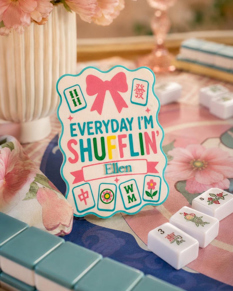 Może przedstawiać: Biały znak z napisem "EVERYDAY I'M SHUFFLIN' Ellen" i grafiką płytek Mahjong. Znak ma r&oacute;żową kokardę i napisy w kolorze r&oacute;żowym, turkusowym i ż&oacute;łtym. Płytki Mahjong są widoczne na pierwszym planie.