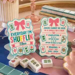 Może przedstawiać: Dwie ozdobne karty do gry w Mahjong z r&oacute;żowymi kokardkami i tekstem. Jedna karta głosi "Everyday I'm Shufflin'", a druga "Jokers cannot be used in a single or a pair". Widocznych jest kilka płytek Mahjong.
