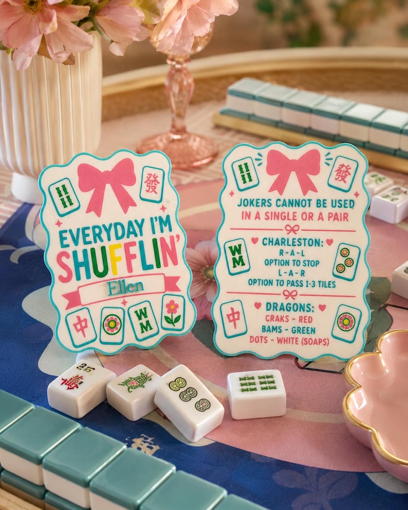 Może przedstawiać: Dwie ozdobne karty do gry w Mahjong z r&oacute;żowymi kokardkami i tekstem. Jedna karta głosi "Everyday I'm Shufflin'", a druga "Jokers cannot be used in a single or a pair". Widocznych jest kilka płytek Mahjong.