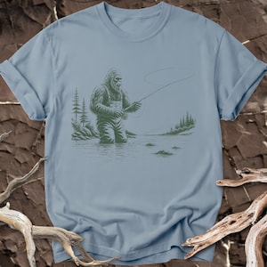 May include: A light blue t-shirt featuring a dark green graphic illustration of a Bigfoot creature wearing overalls and standing in a river while fly fishing with a rod and reel, surrounded by pine trees and a calm water landscape.