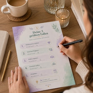 Könnte beinhalten: Eine Person schreibt mit einem Stift auf einem Arbeitsblatt mit dem Titel "Deine 5 größten Fallen". Das Arbeitsblatt hat ein Aquarelldesign und listet fünf potenzielle Fallstricke auf. Eine Tasse Kaffee, eine Kerze und Bücher liegen auf dem Holztisch.