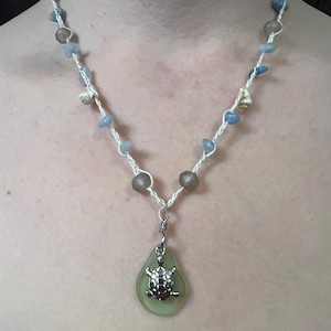 May include: A handmade necklace featuring a sea glass pendant with a silver turtle charm. The necklace is strung with small blue and tan beads, and white cord. The pendant is a light green color.