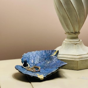 May include: A blue leaf-shaped trinket dish with a textured surface and gold-colored accents along the edges. The dish contains a small silver-toned ring and a gold-toned chain link piece, resting near a tall, off-white, spiraled lamp base.