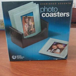 Puede incluir: Una caja para posavasos fotográficos Cherished Accents que incluye un soporte negro y cuatro posavasos de cristal. El embalaje muestra imágenes de personas en los posavasos e incluye el texto "4 glass coasters with holder" y "photo coasters".