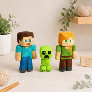 Op de afbeelding: Drie handgemaakte haakfiguren geïnspireerd op een populair videospel: Steve, een Creeper en Alex. Steve draagt een blauw shirt en broek, Alex heeft oranje haar en een groen shirt, en de Creeper is groen. Een houten bordje zegt "handmade with love."