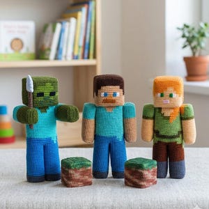 Può includere: Tre bambole all'uncinetto che rappresentano personaggi di Minecraft: uno zombie verde che tiene una spada grigia, una figura dai capelli castani con una maglietta blu e una figura dai capelli arancioni con una maglietta verde, con due blocchi quadrati.