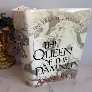 May include: A hardcover book titled "The Queen of the Damned" by Anne Rice. The book is part of "The Vampire Chronicles" series. The cover features a grayscale illustration of a woman's face and the title in bold, black letters.