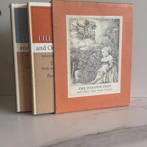 Peut inclure: Trois livres anciens dans un étui. Les livres ont des couvertures bleu clair, orange et beige. L'étui est orange avec une illustration en noir et blanc et le texte "THE JUNIPER TREE and Other Tales from Grimm".