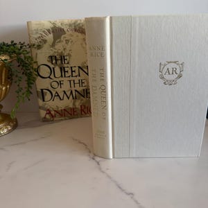 May include: Two hardcover books by Anne Rice are displayed. The book on the left has a gold cover with the title "The Queen of the Damned." The book on the right is white with the initials "AR" in a gold emblem.
