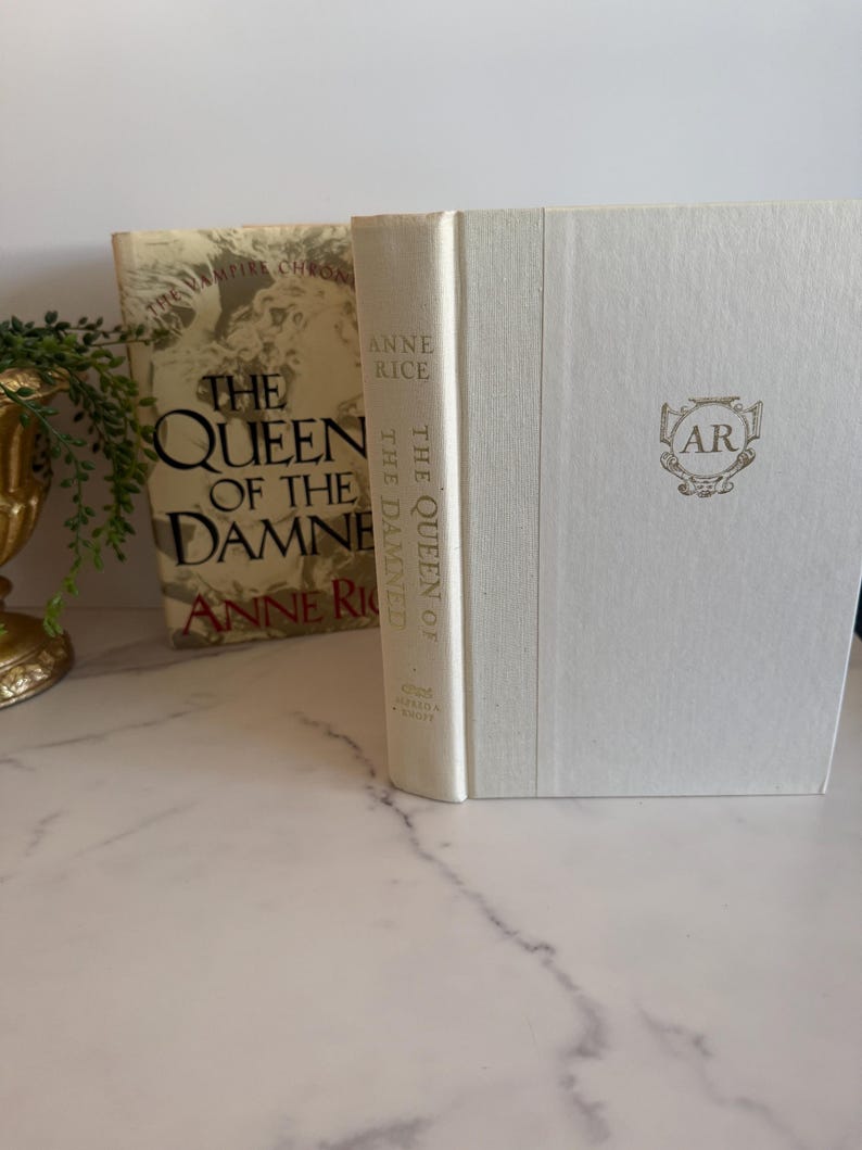 May include: Two hardcover books by Anne Rice are displayed. The book on the left has a gold cover with the title "The Queen of the Damned." The book on the right is white with the initials "AR" in a gold emblem.