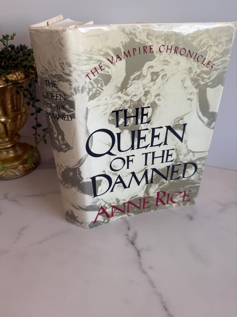 May include: A hardcover book titled "The Queen of the Damned" by Anne Rice. The book is part of "The Vampire Chronicles" series. The cover features a grayscale illustration of a woman's face and the title in bold, black letters.