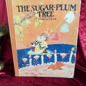 May include: Vintage children's book titled "The Sugar-Plum Tree" by Eugene Field. The cover features a whimsical illustration of a child, gingerbread figures, and colorful candies. The book has an orange and blue color scheme.