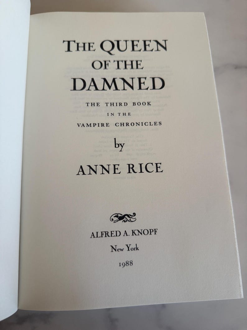 May include: Open book page with the title "THE QUEEN OF THE DAMNED" by Anne Rice. The text is in black font on an off-white page. The book is part of "THE VAMPIRE CHRONICLES" series, published by Alfred A. Knopf in 1988.