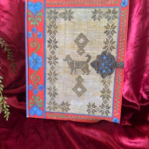 May include: A hardcover book featuring a beige cross-stitched cover with a central dog motif, geometric patterns, and a decorative metal latch. The spine and borders are painted red with blue floral and green vine illustrations.