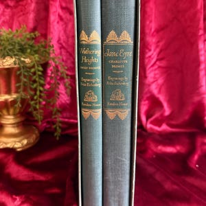 May include: A two-volume book set in a slipcase featuring Wuthering Heights by Emily Bronte and Jane Eyre by Charlotte Bronte. The blue cloth-bound spines display gold lettering, decorative borders, and the text Engravings by Fritz Eichenberg.