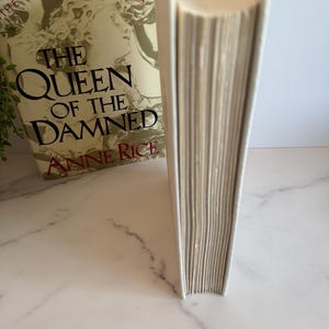 May include: A hardcover book titled "The Queen of the Damned" by Anne Rice. The book has a cream-colored cover and spine, with gold lettering and a portrait of a woman. The book is part of "The Vampire Chronicles" series.