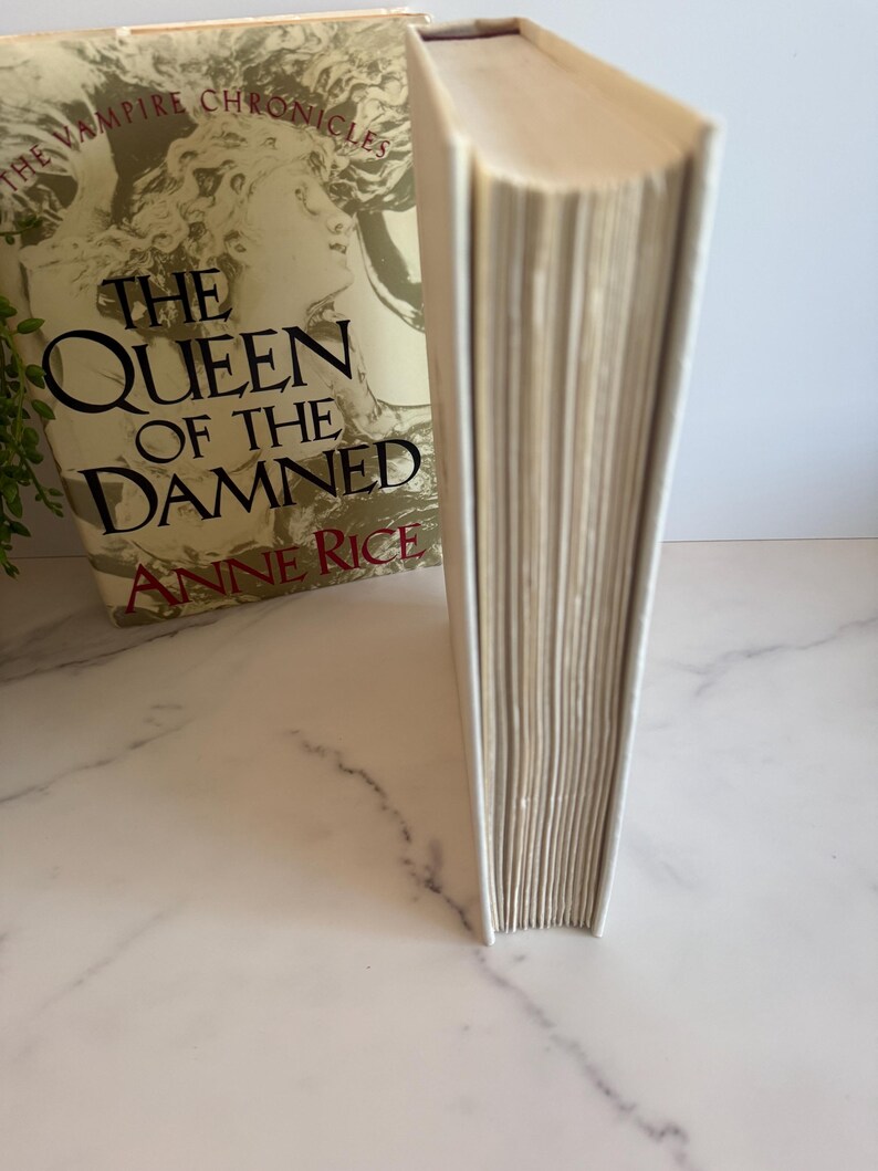 May include: A hardcover book titled "The Queen of the Damned" by Anne Rice. The book has a cream-colored cover and spine, with gold lettering and a portrait of a woman. The book is part of "The Vampire Chronicles" series.