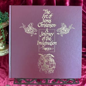 May include: A hardcover book titled "The Art of James Christensen: A Journey of the Imagination" in a deep burgundy color. The cover features gold illustrations of fantastical creatures and text. The book is displayed on a decorative metal stand.