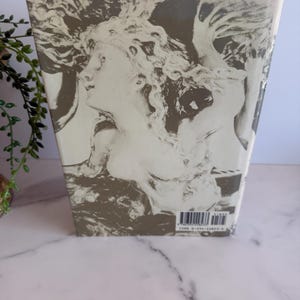 May include: A book with a cover featuring a grayscale illustration of a classical female figure with flowing hair. The artwork is set against a muted background, with the spine visible. The book is wrapped in clear plastic.