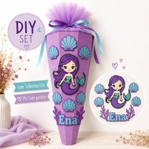 Pode incluir: Um recipiente de presente cônico roxo com uma ilustração de sereia, recortes de conchas em azul-petróleo e o nome Ena em letras da mesma cor. O topo é finalizado com tule roxo e uma fita azul. Texto: DIY SET, zum Selberbasteln, Mit Liebe gestaltet.
