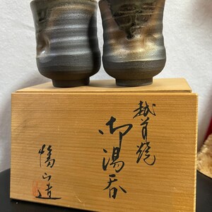 以下が含まれることがあります： テクスチャ加工された濃いグレーと茶色の釉薬が施された、手作りのセラミックティーカップ2個。 カップは、日本の書道が施された木箱の上に置かれています。 カップはユニークで有機的な形をしています。
