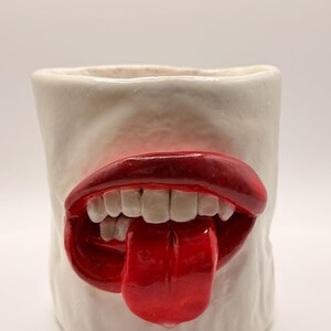 Könnte beinhalten: Ein weißer Keramikbecher mit einem Design aus rotem Mund und Zunge. Der offene Mund hat weiße Zähne und eine glänzende rote Zunge, die herausragt. Der Becher hat eine glatte, matte Oberfläche.
