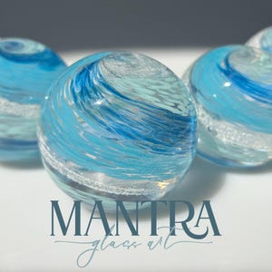 May include: Five spherical glass paperweights featuring swirling patterns of light blue, dark blue, and white colours. The glass spheres have a smooth, polished surface with internal bubbles and textures. The text MANTRA glass art is printed below.