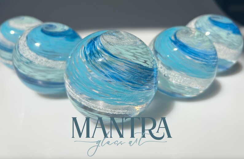 May include: Five spherical glass paperweights featuring swirling patterns of light blue, dark blue, and white colours. The glass spheres have a smooth, polished surface with internal bubbles and textures. The text MANTRA glass art is printed below.