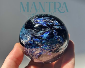 May include: A spherical glass paperweight featuring a deep black base with swirling patterns of metallic blue, silver, and iridescent glitter. The surface is smooth and reflective. The text MANTRA glass art is visible in blue above the sphere.