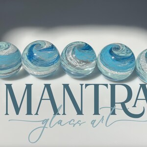 May include: Five spherical glass paperweights arranged in a horizontal row, each featuring swirling patterns of light blue, dark blue, and white with embedded silver glitter. The text MANTRA glass art is printed in dark blue below the spheres.