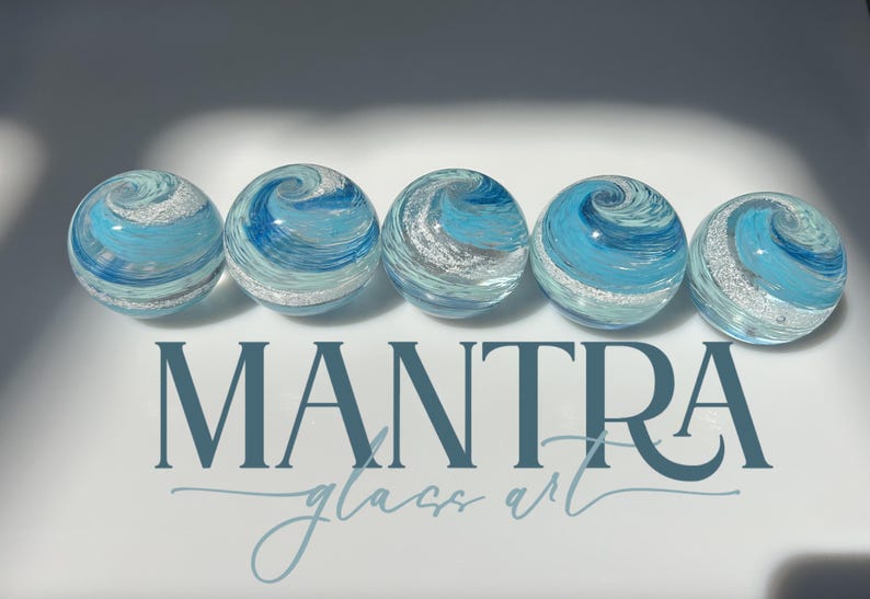 May include: Five spherical glass paperweights arranged in a horizontal row, each featuring swirling patterns of light blue, dark blue, and white with embedded silver glitter. The text MANTRA glass art is printed in dark blue below the spheres.