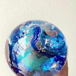 May include: A glass orb with blue, white, and iridescent glitter swirls inside. The orb is clear and has a smooth, round shape.