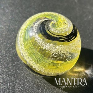 May include: A clear glass marble with a yellow and black spiral design. The marble has a swirl pattern with white speckles throughout. The text "MANTRA glass art" is visible at the bottom.