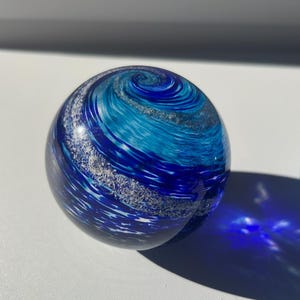 May include: A spherical glass paperweight with a swirling blue and white design. The glass orb has a dark blue base with lighter blue and white accents, creating a dynamic, ocean-like pattern. The paperweight is smooth and reflective, casting a blue shadow.