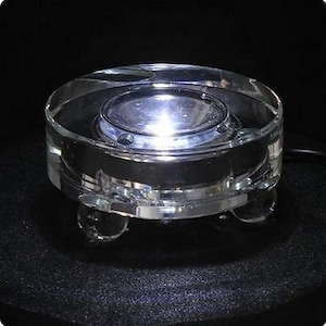 May include: A clear, round, glass coaster with a built-in light. The coaster has a raised center and sits on three small, clear, spherical feet. A black cord extends from the side. The coaster is designed to hold a small object.