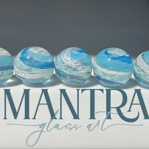 May include: Five spherical glass paperweights arranged in a row, each featuring swirling patterns of light blue, dark blue, and white with embedded silver glitter. The text MANTRA glass art is printed in dark blue below the glass spheres.