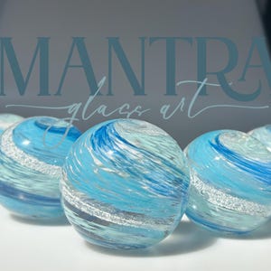 May include: Five spherical glass paperweights arranged in a row, each featuring swirling patterns of light blue, dark blue, and white glass with a distinct band of shimmering silver glitter embedded within the clear, polished glass surface.