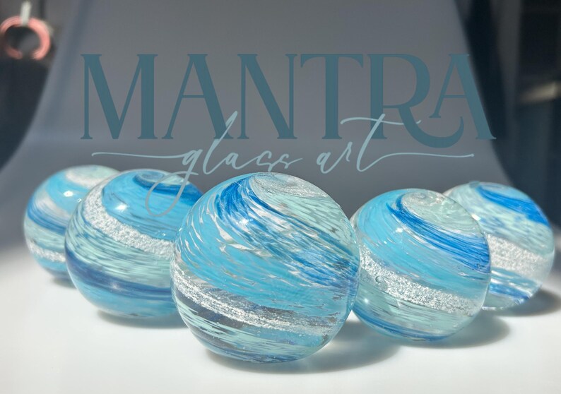 May include: Five spherical glass paperweights arranged in a row, each featuring swirling patterns of light blue, dark blue, and white glass with a distinct band of shimmering silver glitter embedded within the clear, polished glass surface.