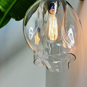 May include: A clear glass light fixture with a wavy, organic shape. The fixture has a single light bulb inside, which is turned on and glowing.