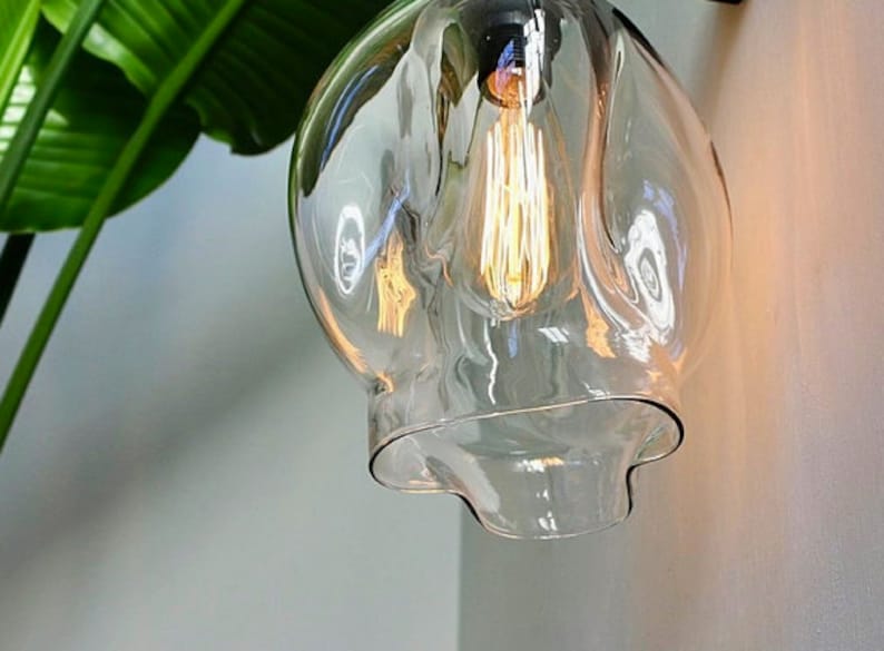 May include: A clear glass light fixture with a wavy, organic shape. The fixture has a single light bulb inside, which is turned on and glowing.